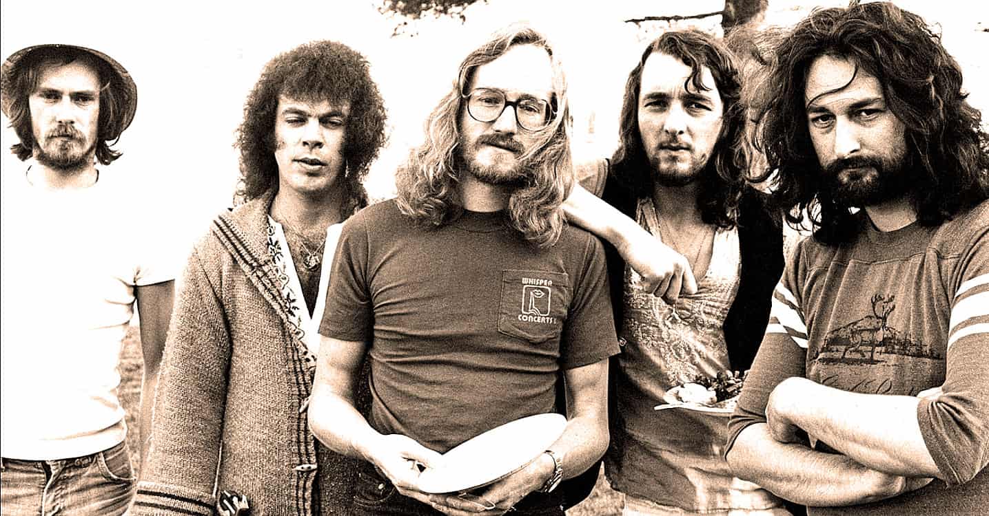 A vintage black-and-white photo of a 1970s rock band outdoors, showcasing musicians with long hair and casual attire, emphasizing music history and classic rock.