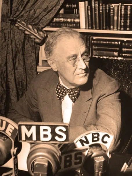 Vintage black and white photograph of a distinguished man in glasses, sitting at a desk with microphones labeled MBS, NBC, and CBS, surrounded by books and a curtain, representing historical radio broadcasting.
