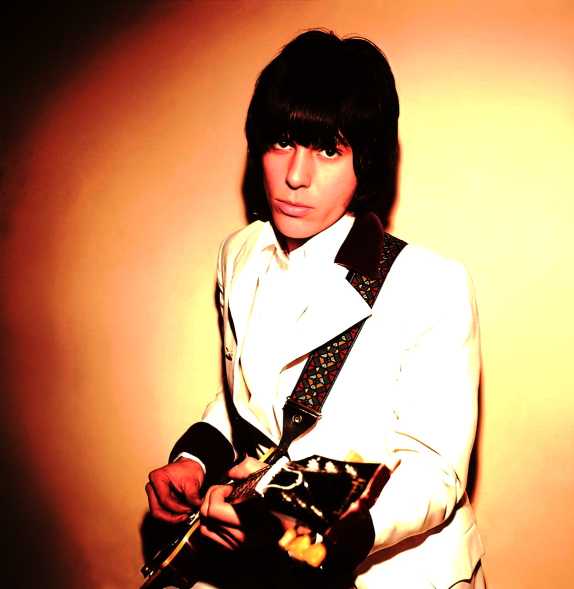 - A young male musician with dark hair holding an electric guitar, wearing a white suit with black collar details, against a warm gradient background.