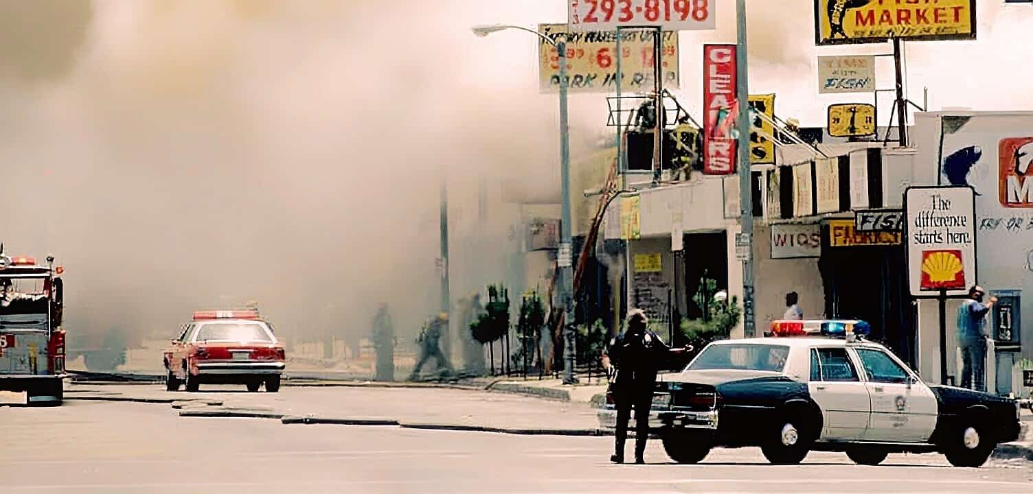 Fire emergency response scene, smoke billowing from building with police and fire vehicles present, urban street with commercial signs, illustrating historic news coverage and public safety events.