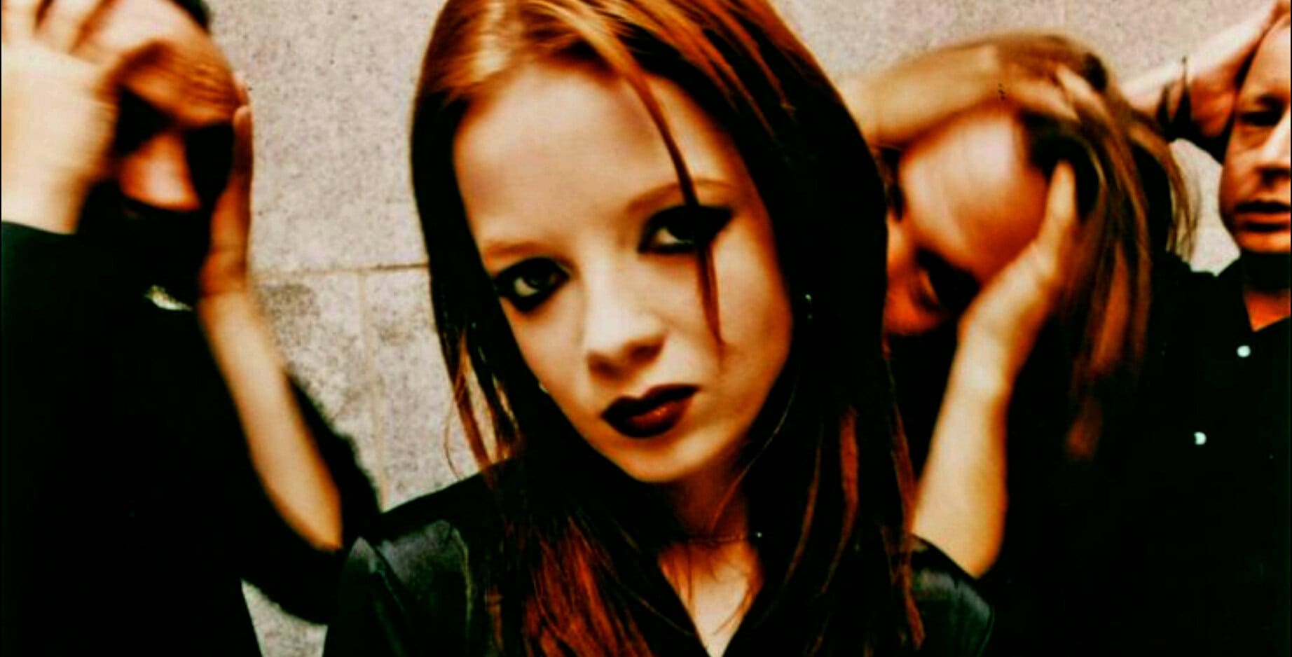 Dark-haired woman with intense makeup and gothic style, posing against a concrete wall, conveying a rebellious music vibe, linked to news, history, and music archives on Past Daily.