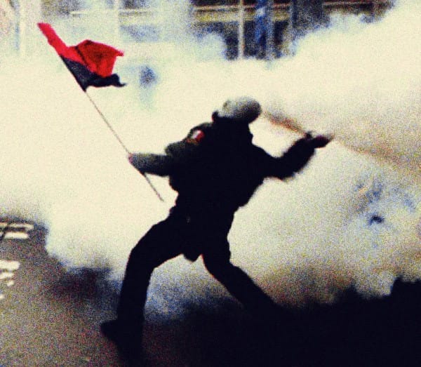 Police officer in riot gear throwing tear gas during protest, protester holding red and black flag, civil unrest, social movement, law enforcement confrontation, chaos in public space.