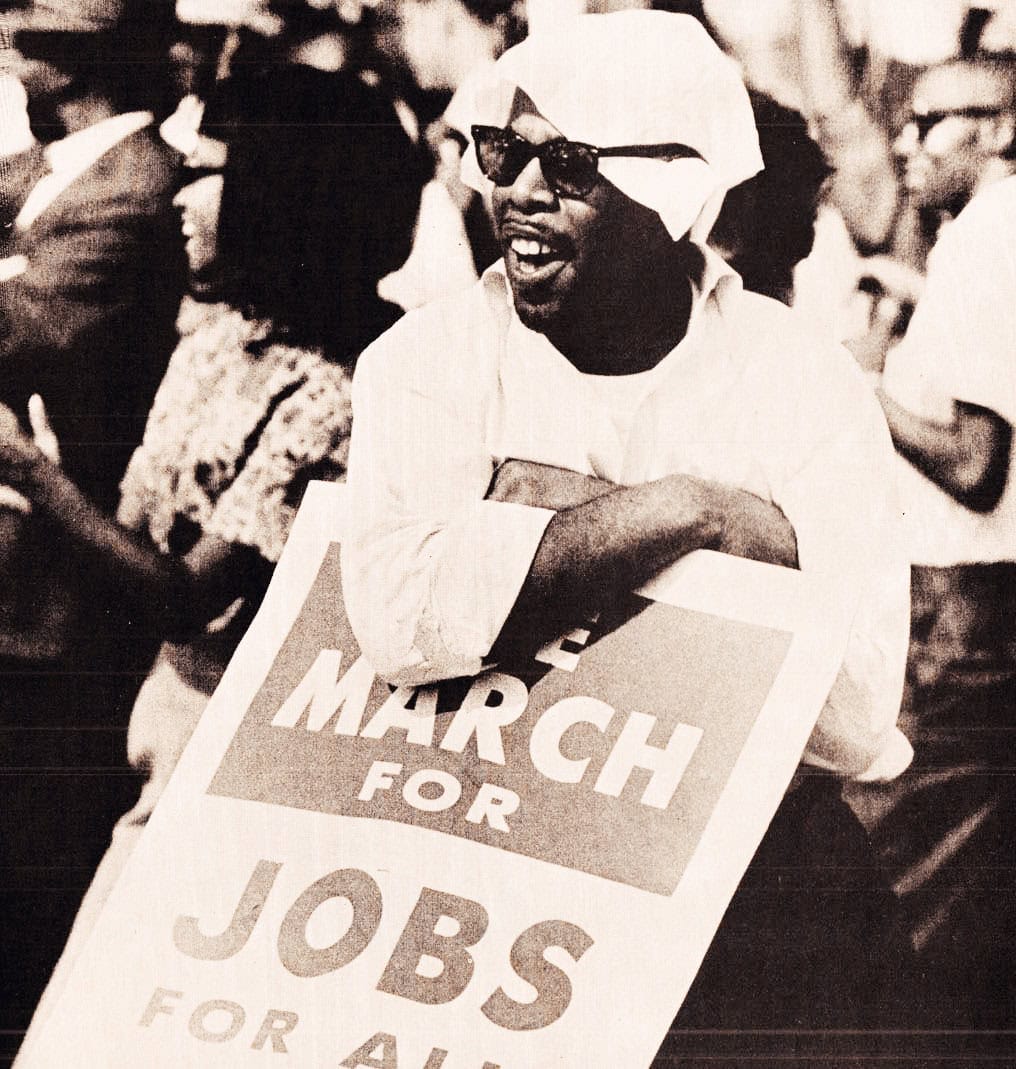 Protest marcher holding a "March for Jobs for All" sign at a historic demonstration, emphasizing social justice, economic equality, and civil rights; iconic black-and-white photo capturing activism.
