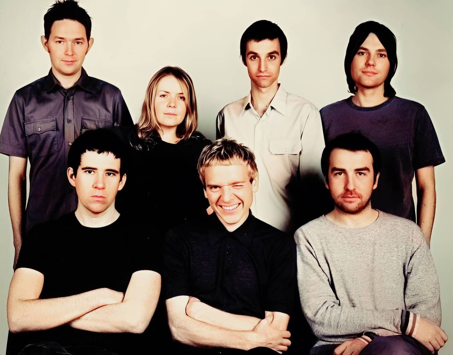 A black and white indie rock band group portrait of seven young adults, with some crossing their arms and one person winking, representing music and band culture.