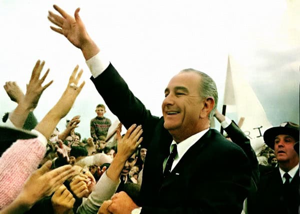 A happy man in a suit waving to a crowd at a political rally or event with supporters reaching out.