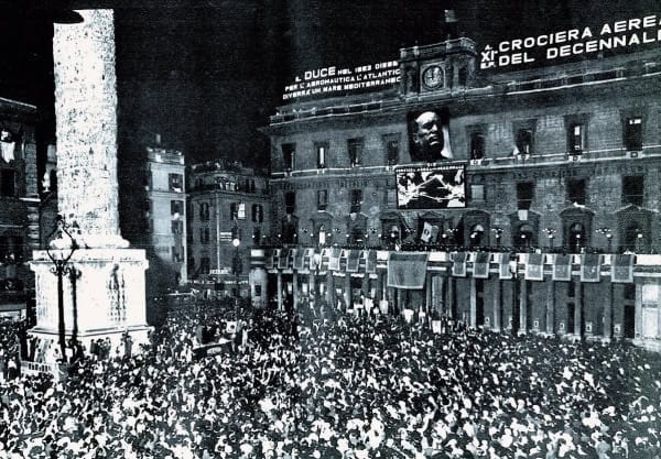 Massive crowd gathered in front of a historic building during a significant event, capturing a lively atmosphere with banners and digital screens, related to news, history, and music archives.