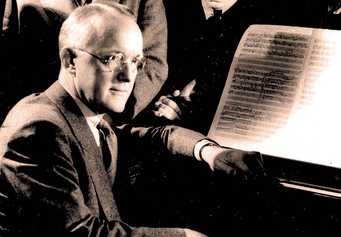 A black and white image of a man in a suit playing the piano with sheet music, capturing a historical music recording session for the Past Daily sound archive.