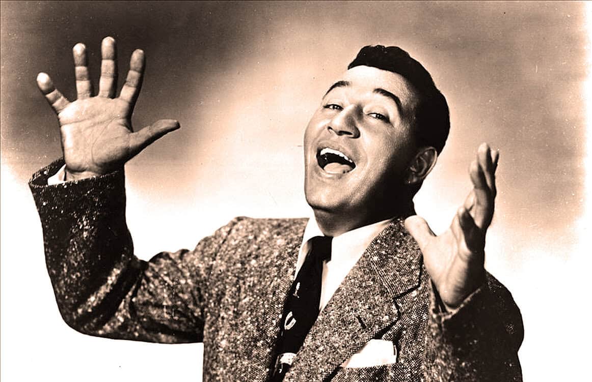 Vintage black and white photo of a lively male singer performing with expressive gestures, wearing a glittery jacket and tie, capturing classic music and entertainment history from the past.