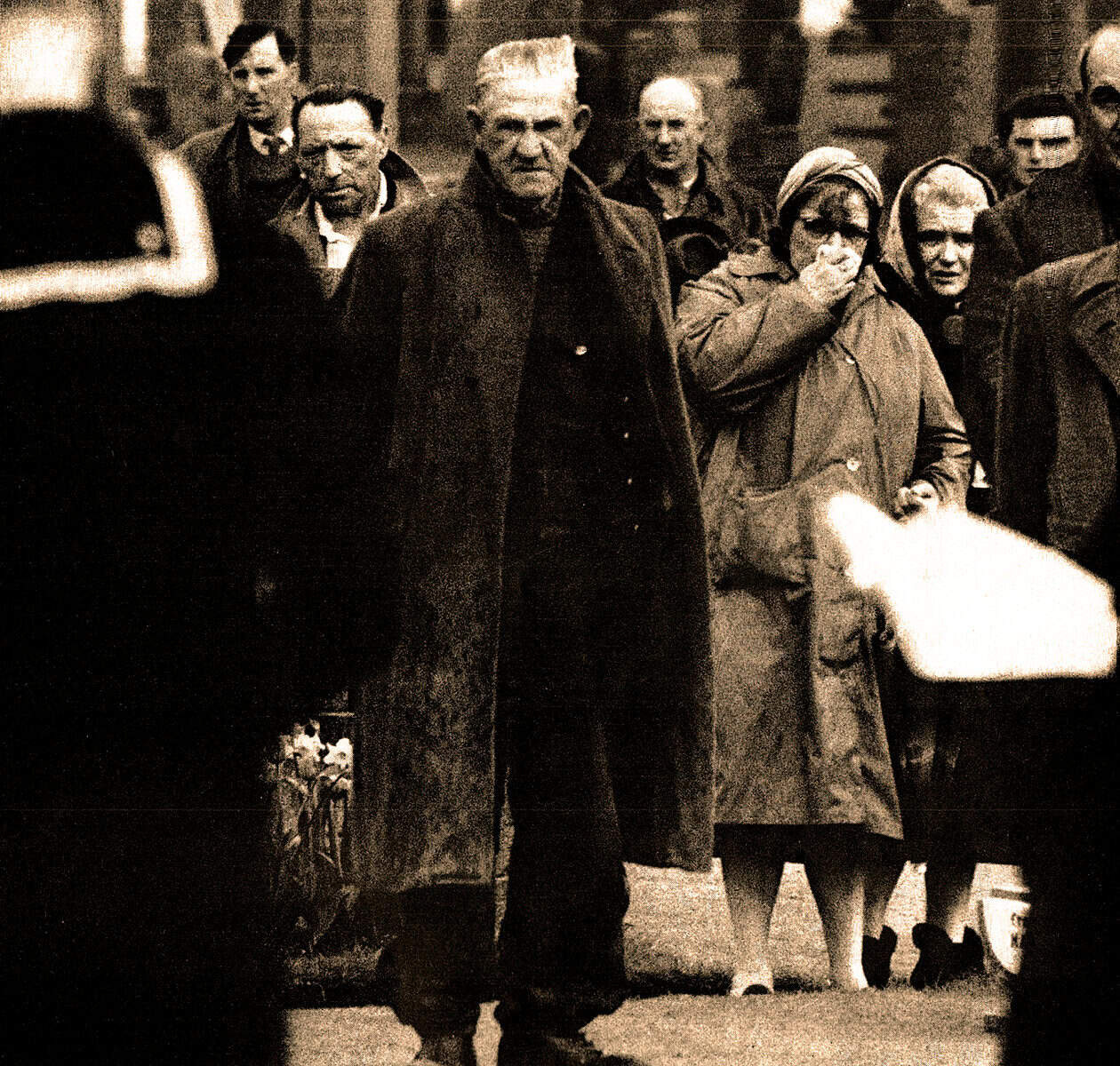 People gathered outdoors, some wearing coats and head coverings, with a focus on an elderly man and woman, capturing a historical moment related to news, history, and music archives.