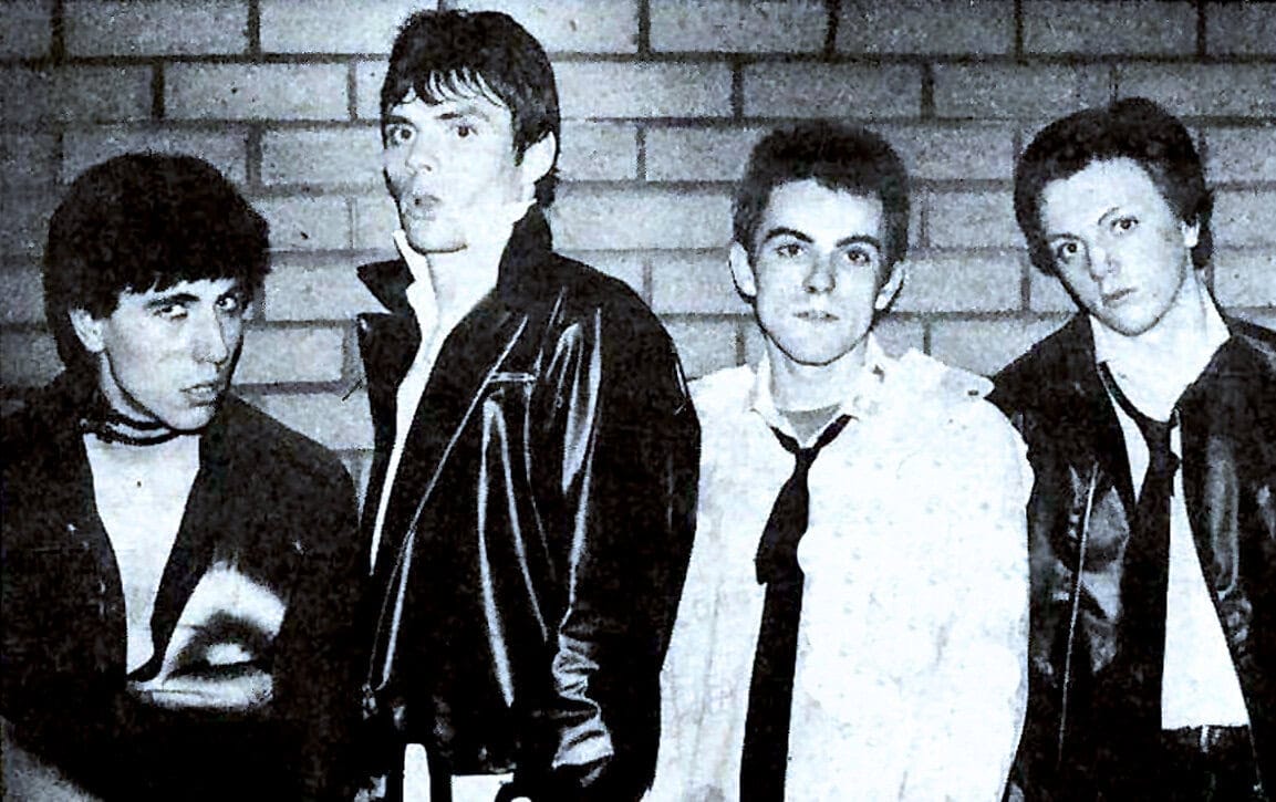 Loud-backed rock band members wearing leather jackets posing in front of brick wall for music history and news archive.