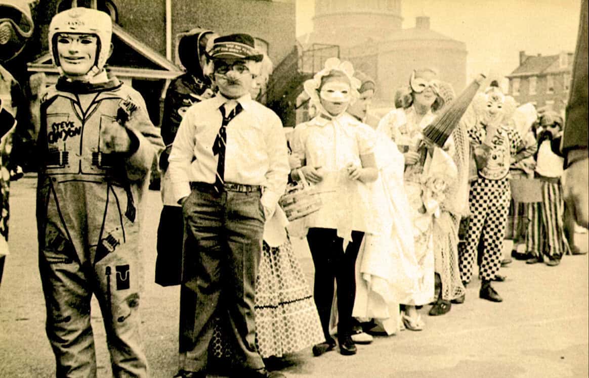 People dressed in vintage costumes and masks during a historical or cultural event, possibly a parade or festival, showcasing nostalgia and community traditions.