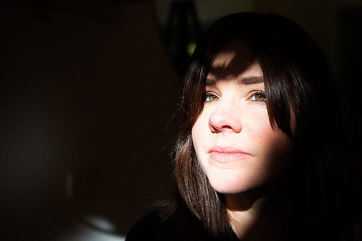 Beautiful young woman with light skin and brown hair, illuminated by sunlight through window blinds, showcasing a contemplative expression.