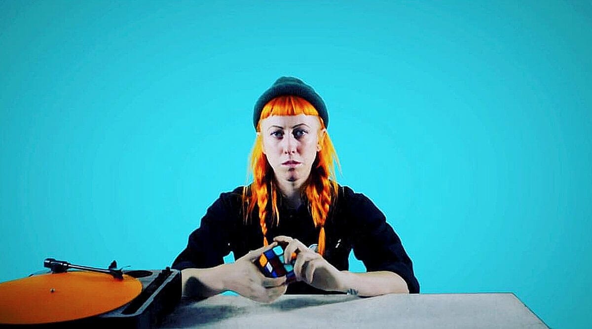 Past Daily archive, news, history, and music memorabilia featuring a young woman with vibrant orange hair using a Rubik's Cube with a record player on a blue background.