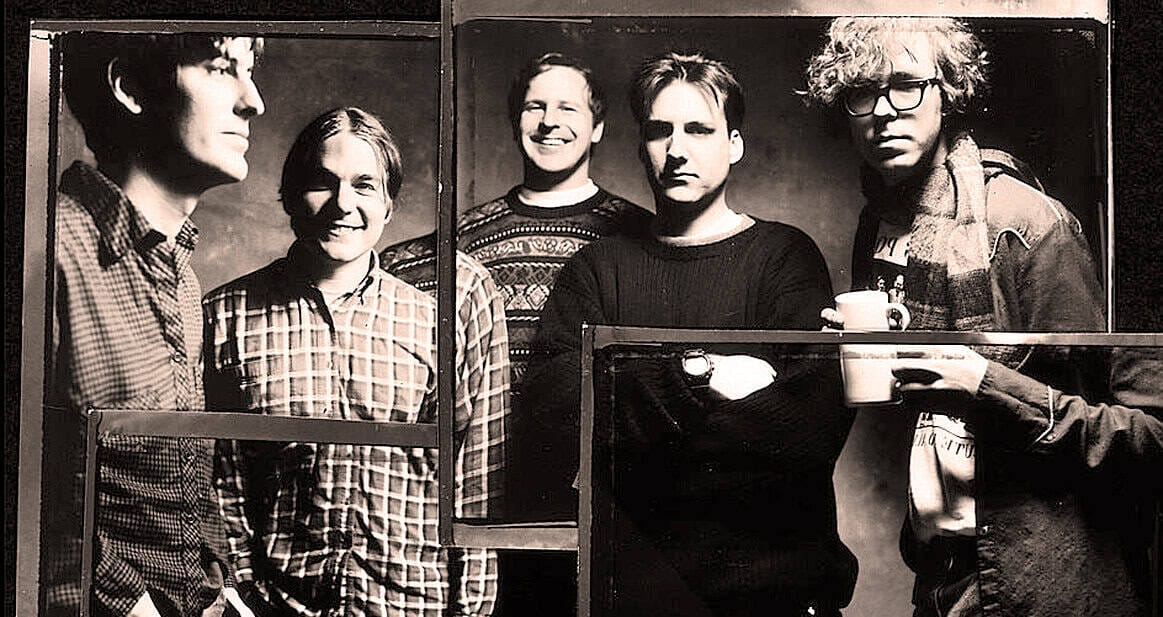 "Black and white photo of the band Radiohead, known for alternative rock and experimental music, posing with crossed arms and holding coffee cups, highlighting their artistic style and influence in music history.".