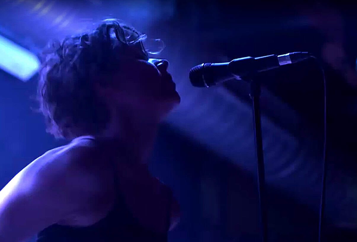 Young woman singing into microphone in a dimly lit stage with blue lighting, capturing live music performance for sound archive of news, history, and music recordings.