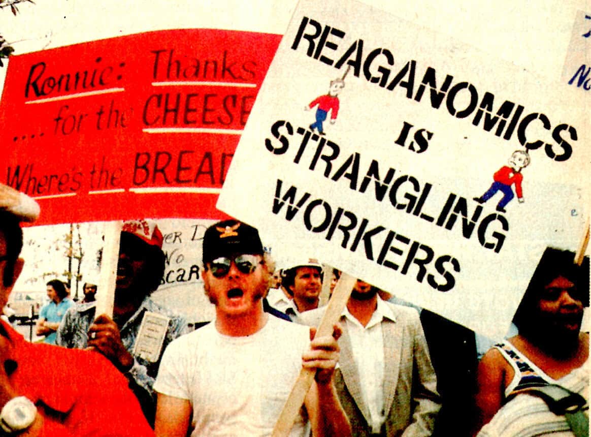 Protesters holding signs about Reaganomics and worker rights during a labor demonstration, emphasizing economic history and social activism.