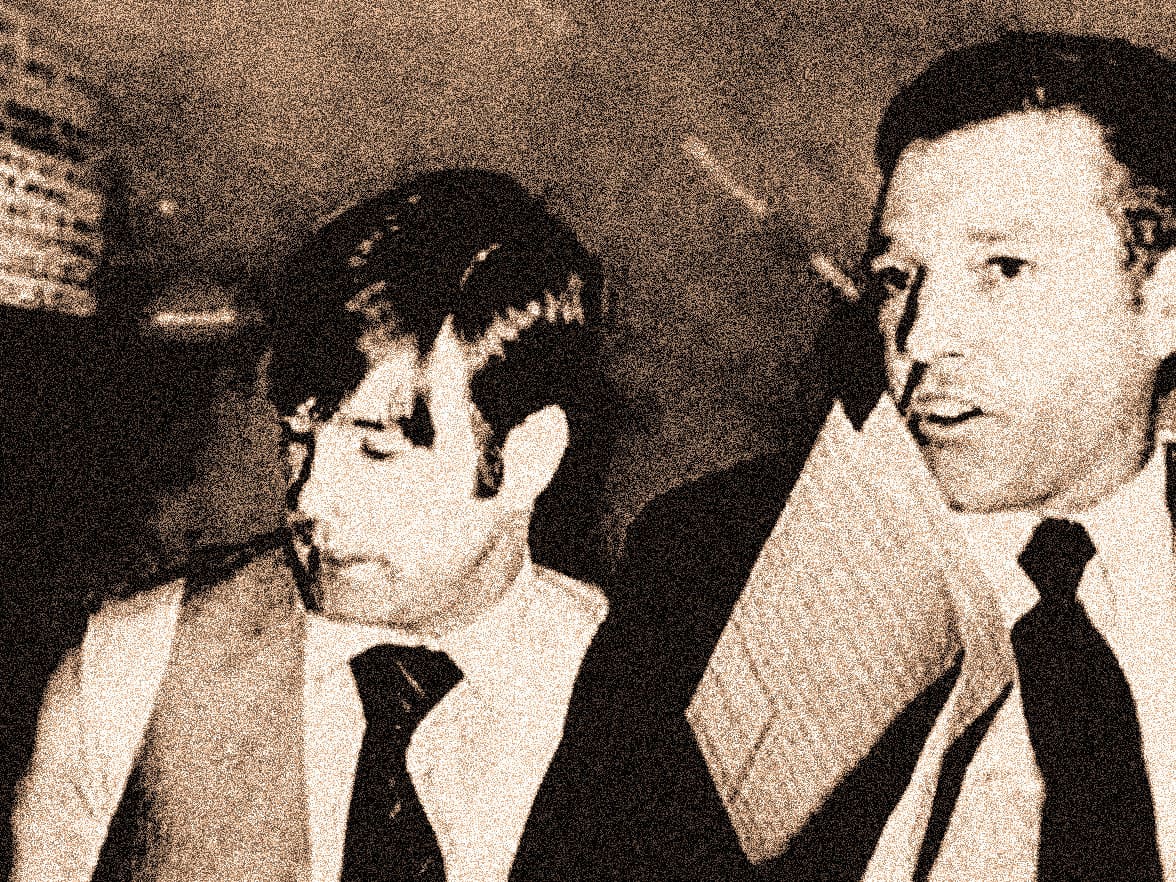 Vintage black-and-white photo of two men in suits and ties engaged in conversation, capturing a moment of historical news and music archive content from Past Daily.