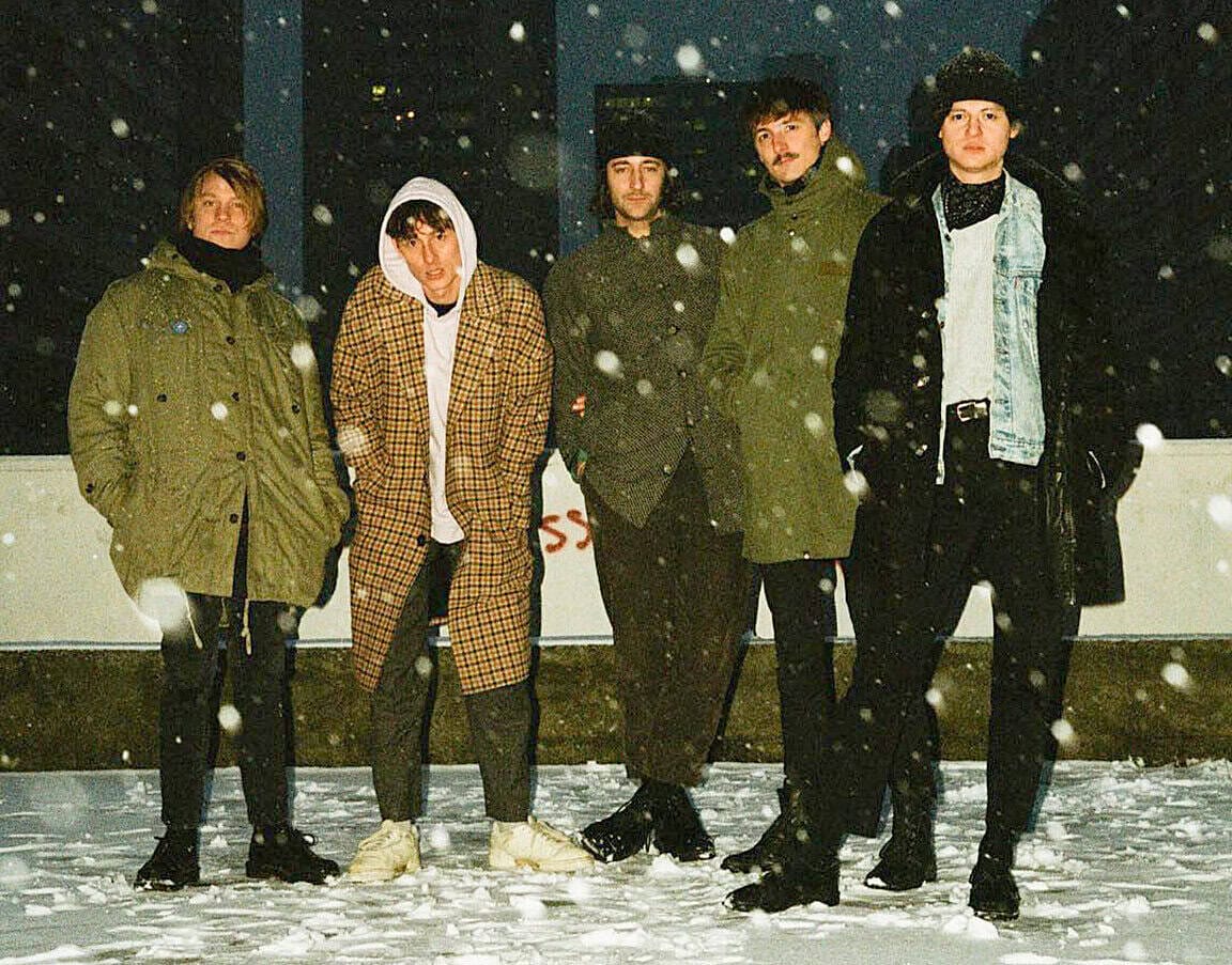 Snowy night outdoor music band members standing together in winter weather, rock band style photo, snow falling, casual clothing, harbor or outdoor concert setting, music history image, music archive, Past Daily music collection.
