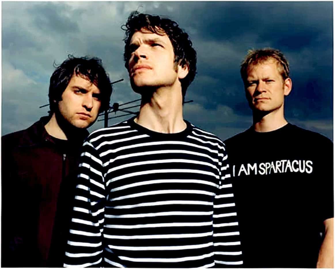 Band of three men outdoors with stormy sky background, one wearing a striped shirt, focusing on music history and classic rock plays, part of a sound archive.