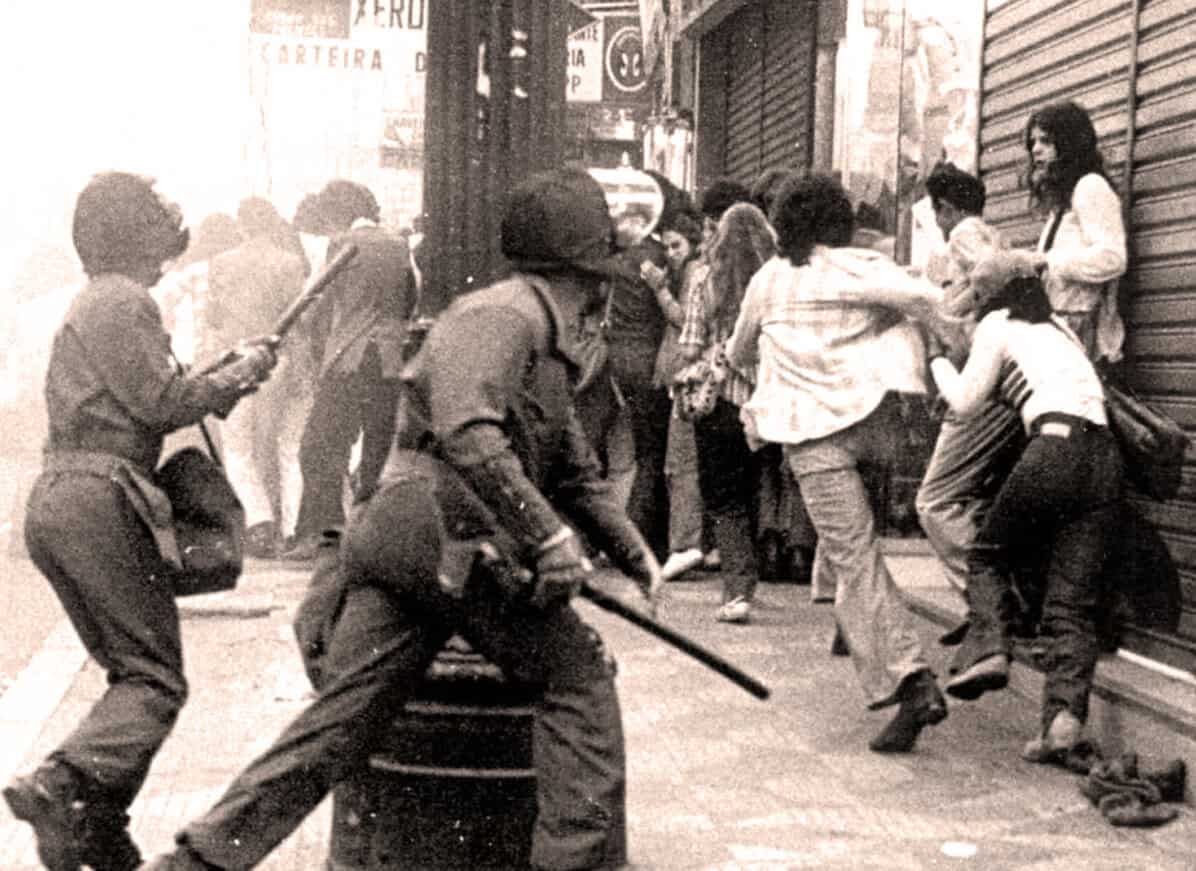 Riot police clash with protesters in urban street scene during civil unrest.
