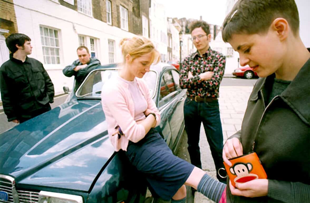 A group of young adults gathered around a vintage car on a city street, engaging in a casual conversation, showcasing city life, youth culture, and social interaction, with a background of European-style architecture.
