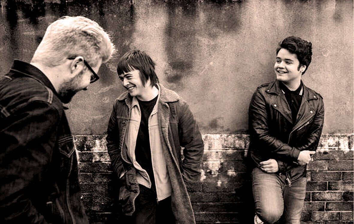A black and white photo of three people laughing and talking outdoors, showcasing casual fashion, friendship, and social interaction; ideal for music, news, and history archive images.