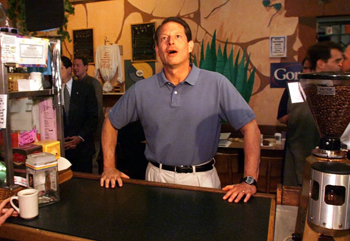 Man speaking behind a coffee shop counter, capturing a lively scene at a café or restaurant, with vibrant background artwork and barista equipment, emphasizing community and social interaction.