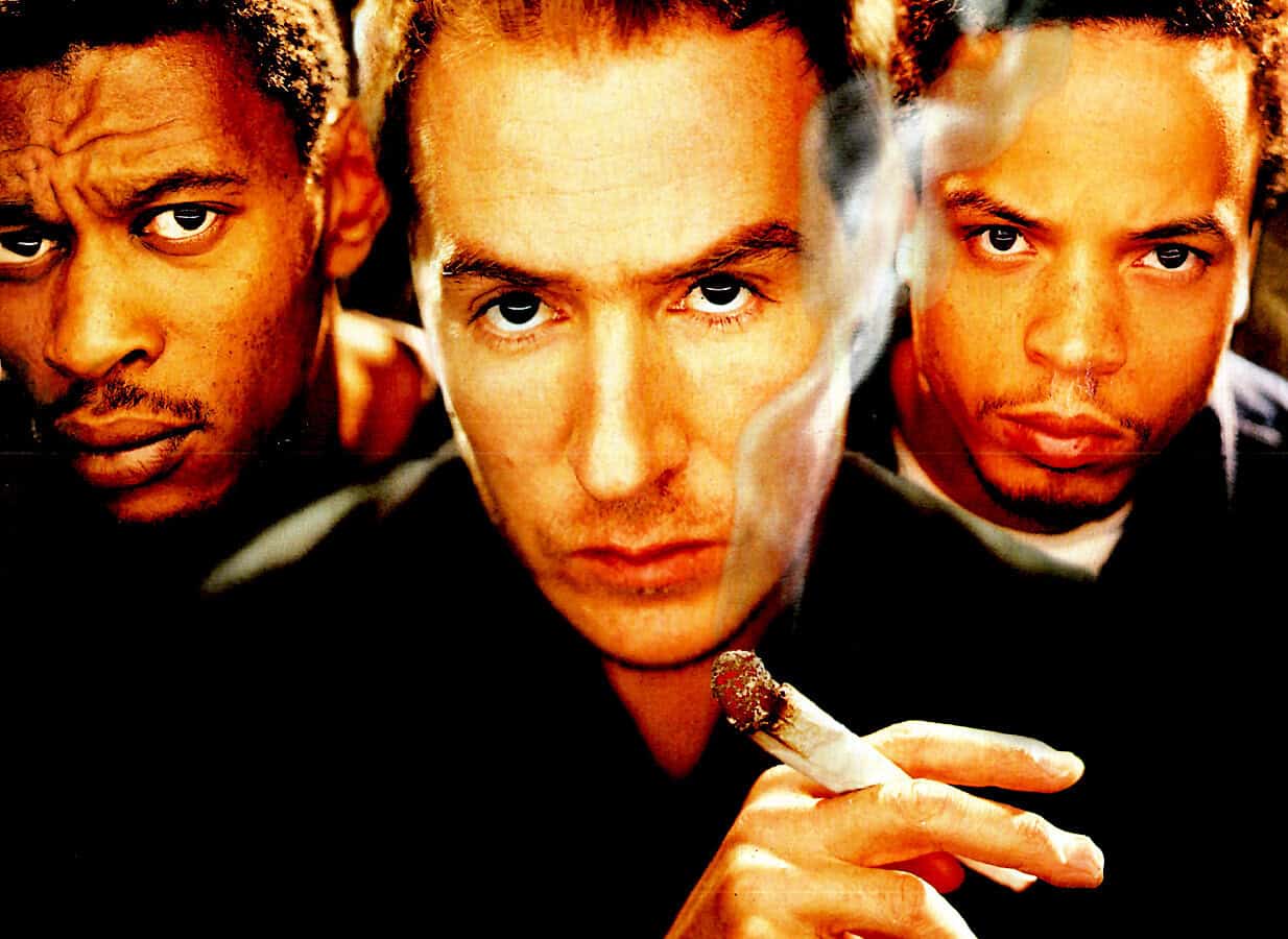 Harsh lighting portrait of three young men with intense expressions, one smoking a cigarette, reflective of urban music culture and modern style, ideal for music history and news archives.