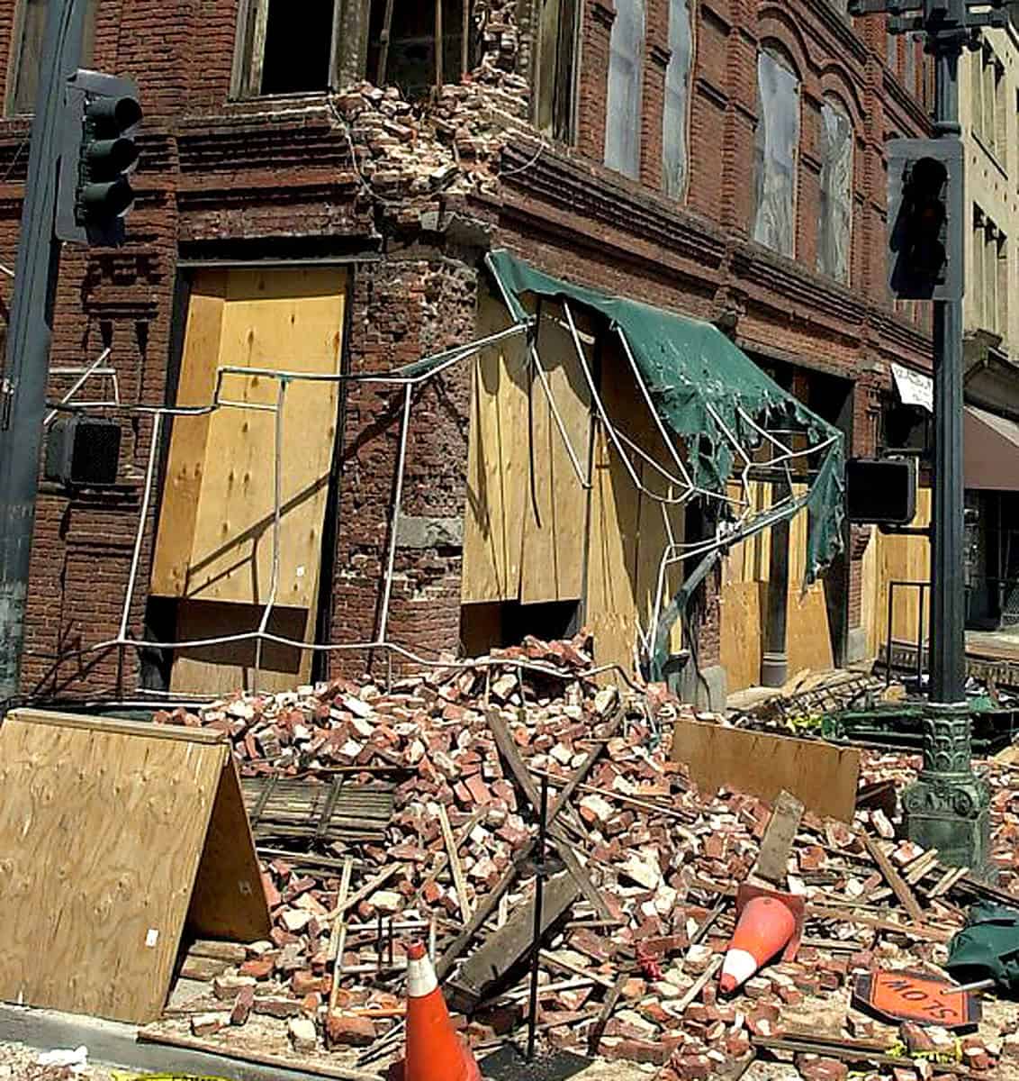 Damaged brick building after collapse, with debris and fallen bricks on the street, emergency cones, and a toppled green awning, highlighting urban destruction scenes.