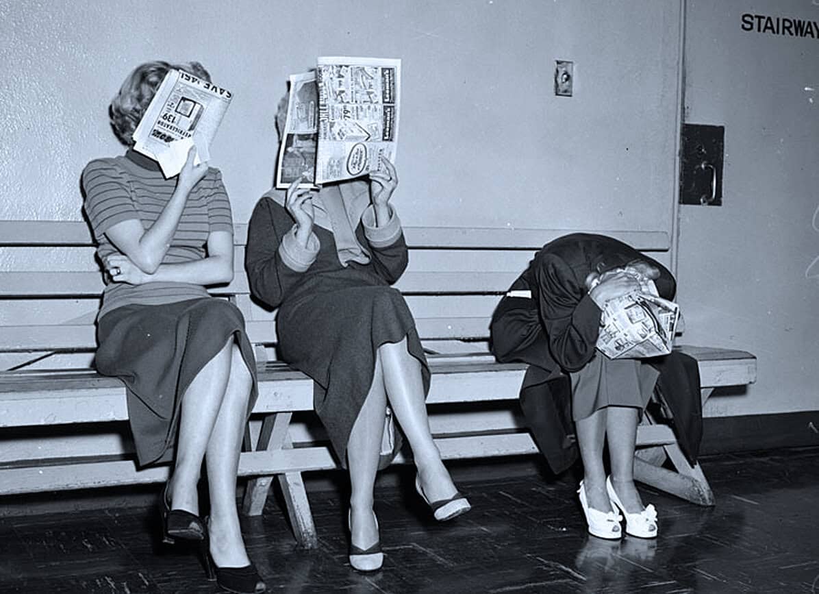 Children sitting on a bench covering their faces with newspapers, in a vintage black and white photo, representing social history, childhood, and newspapers from the past.