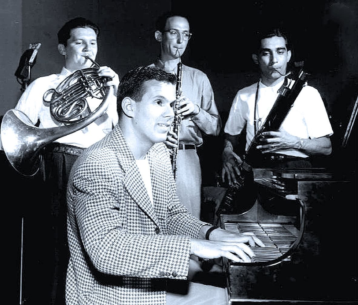 A black and white photo of four jazz musicians playing instruments including piano, trumpet, clarinet, and bass saxophone, capturing a jazz performance from the mid-20th century.