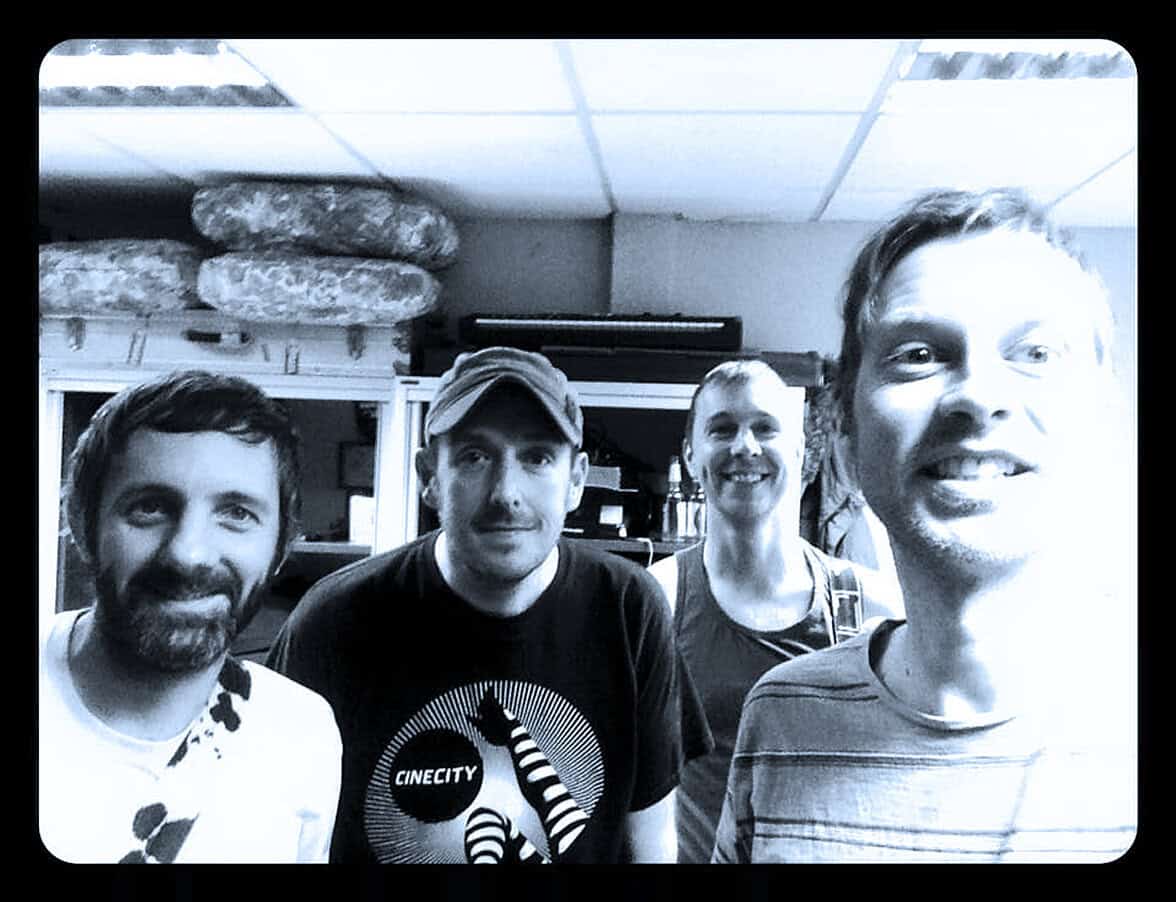 Men wearing casual clothing in recording studio with soundproof padding and musical equipment.