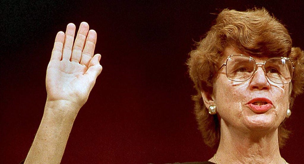 "ALT text: Woman with short curly hair and glasses raising her right hand in a historical or official context, wearing pearl earrings.