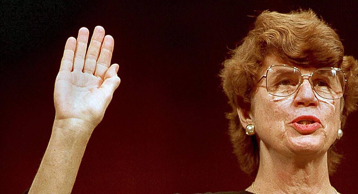 "ALT text: Woman with short curly hair and glasses raising her right hand in a historical or official context, wearing pearl earrings.