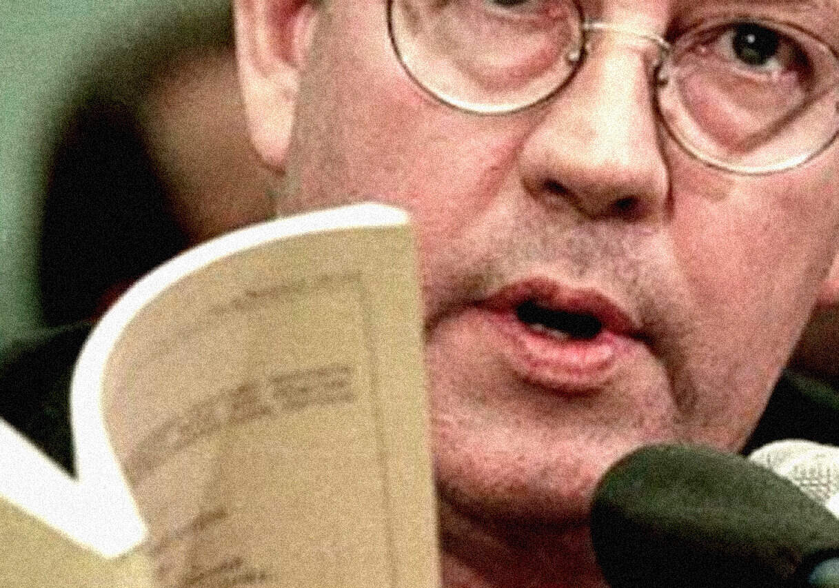 Close-up of a man wearing glasses speaking into a microphone, with a coffee mug partially visible in the foreground. The image captures a moment of speech or interview, emphasizing vocal and media engagement.