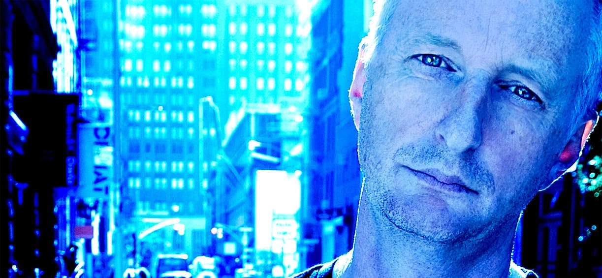 A vibrant city street with bright blue lighting and tall buildings, featuring a close-up of a man with a contemplative expression, representing urban life and urban culture.
