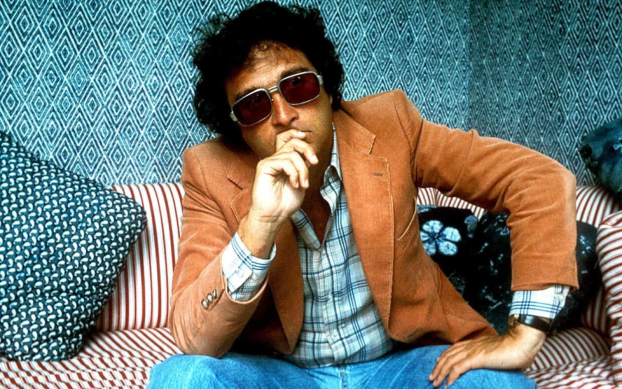 Retro man wearing sunglasses and a brown blazer sitting on a striped couch with patterned pillows, evoking 1970s style, history, music, and news archival themes.