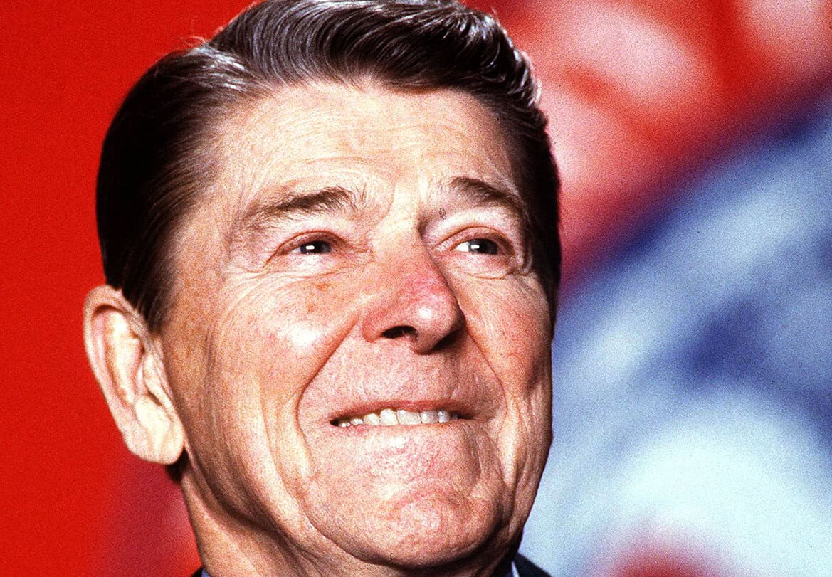 Senior man smiling at a political event, symbolizing history and news coverage, with a blurred background hinting at American patriotism and music archives.