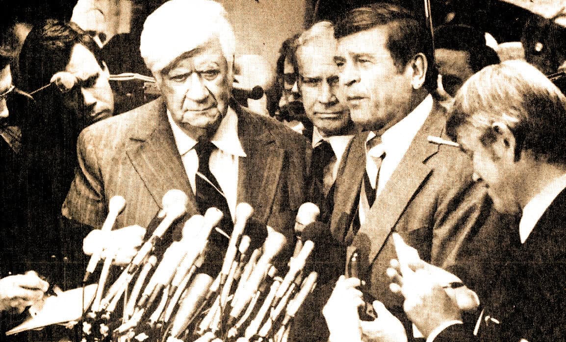 A vintage black and white photo of a group of politicians and journalists at a press conference with multiple microphones, capturing a historical moment in news, politics, and media history.