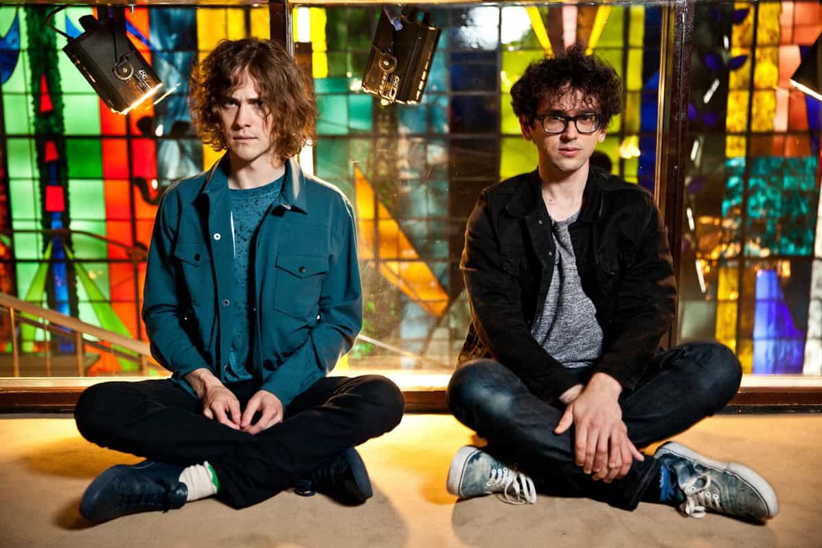 Young men sitting cross-legged in front of a colorful stained glass window, highlighting music, youth culture, and creative expression.