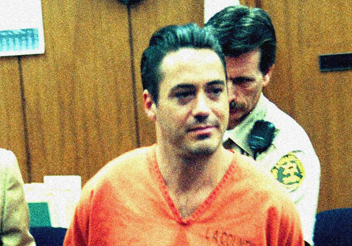 Man in orange prison jumpsuit escorted by sheriff deputy, in a courtroom or detention setting.