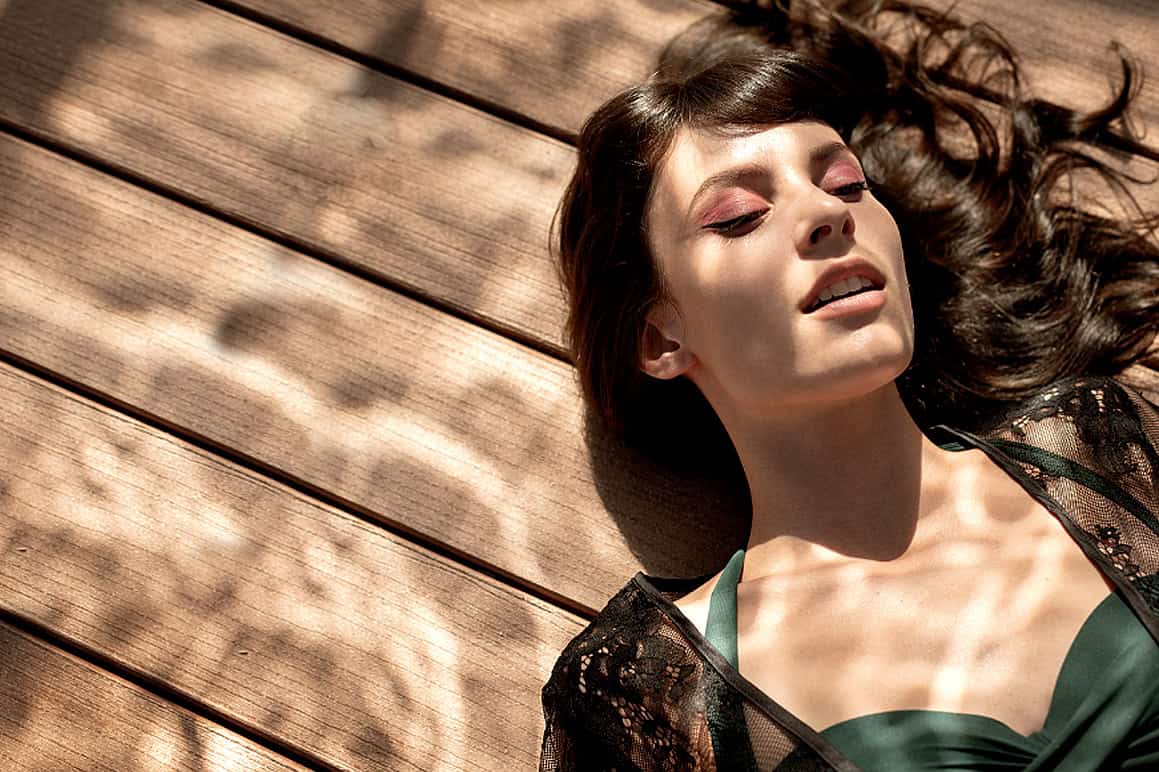 Beautiful woman lying on wooden deck with sunlight and shadows, showcasing fashion photography, outdoor leisure, and stylish appeal.