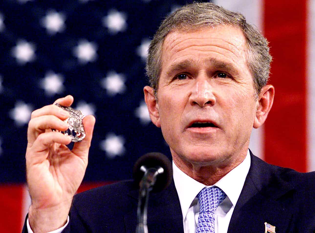 Historical political figure holding a silver badge, speaking at a press conference with American flag background, related to US politics, government, leadership, and history.