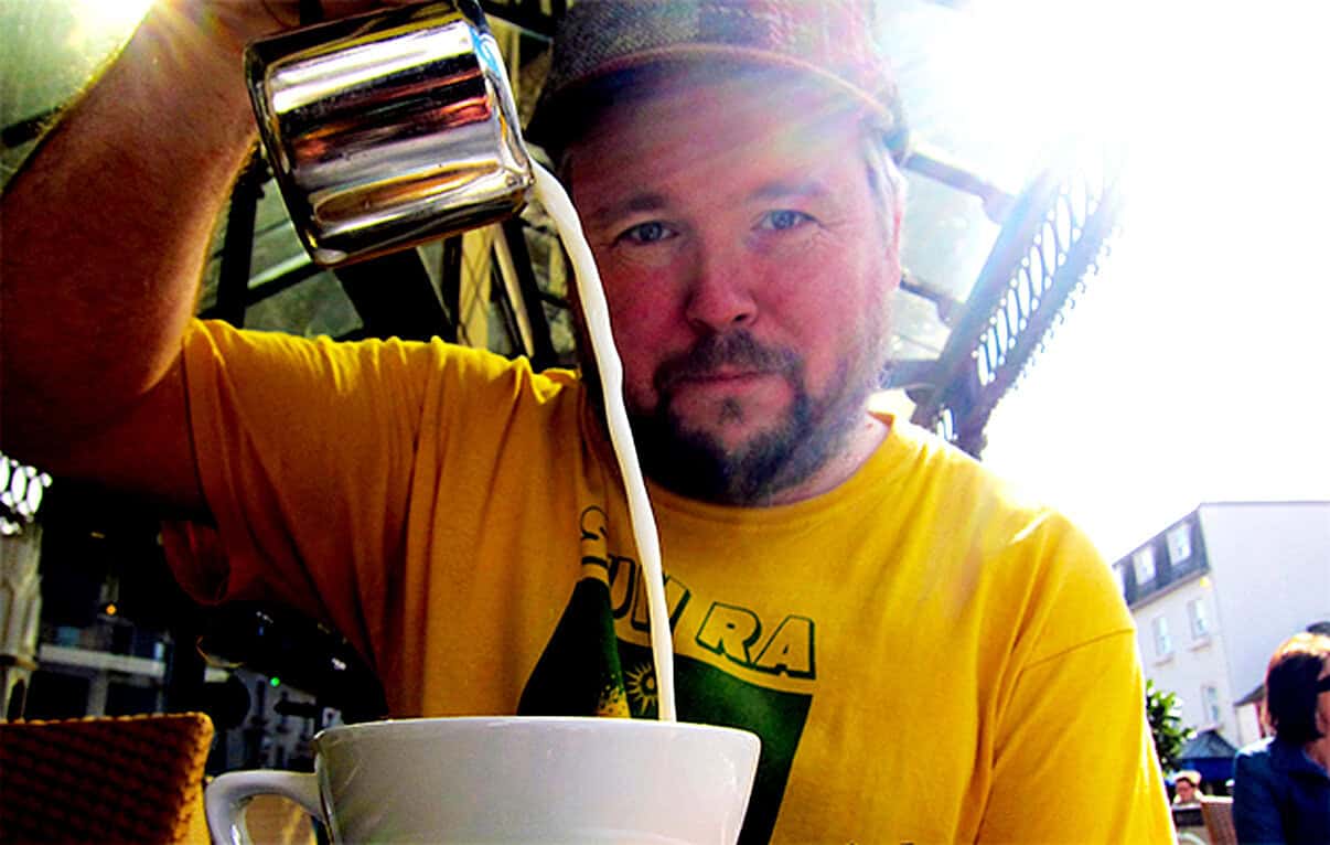Delicious coffee being poured into a white mug outdoors, person in yellow shirt, sunny day, casual dining, urban cafe atmosphere, morning beverage, relaxed lifestyle, Past Daily archive image, vibrant and inviting scene, promoting sound archive of news, history, and music.