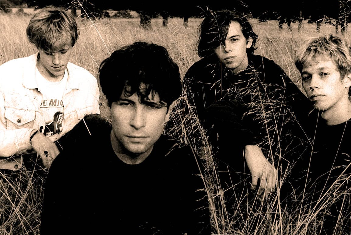 Vintage photo of a rock band in a grassy field, featuring four young male musicians, capturing the essence of 1980s alternative music scenes, perfect for music archives and history enthusiasts.