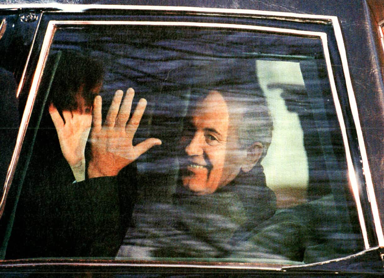 A man waving from inside a vehicle, smiling and greeting outdoors, captured through a car window.