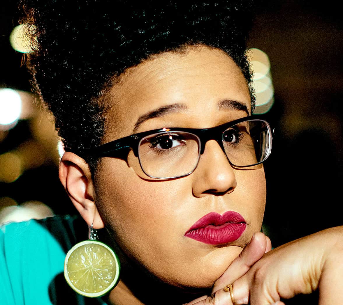 Vibrant portrait of a woman with stylish black glasses, bold makeup, and lime-green fruit earrings, capturing her thoughtful expression in a night-time urban setting for nostalgic audio and music history.