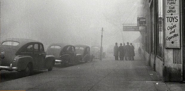 A historic black and white photograph of a foggy urban street scene with vintage cars and pedestrians, capturing moments from mid-20th-century news, history, and music archives.