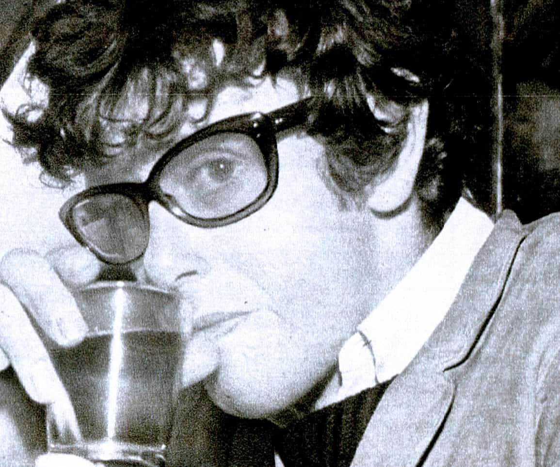 Close-up black and white photo of a young man with curly hair, wearing glasses, holding a drink with a straw, reflecting vintage style and mood, representing music, news, and history archives.