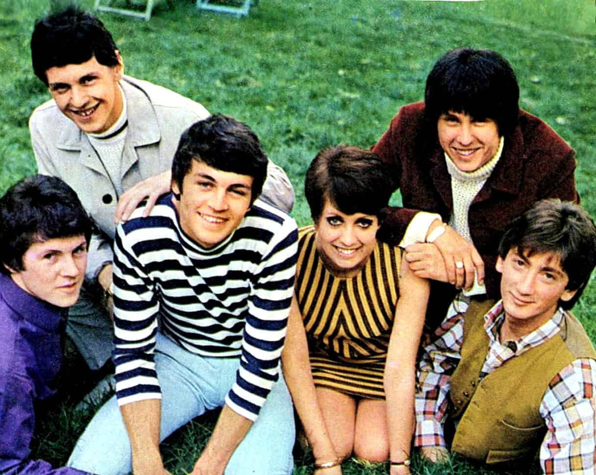Group of young people smiling outdoors, 1960s, casual fashion, pop culture, friendship, vintage photograph, nostalgic, music and entertainment, social gathering, colorful clothing, lively atmosphere, memory lane, classic era of youth culture, timeless moments.